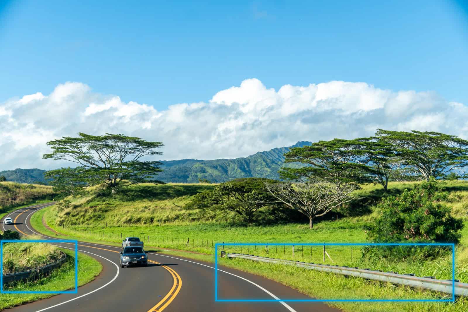 A car drives along a winding road bordered by grass, trees, and mountains under a partly cloudy sky. Guardrails line sections of the roadside.