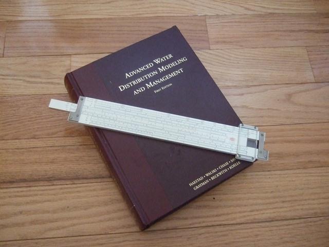 A slide rule resting on top of a textbook titled "advanced water distribution modeling history and management" on a wooden floor.