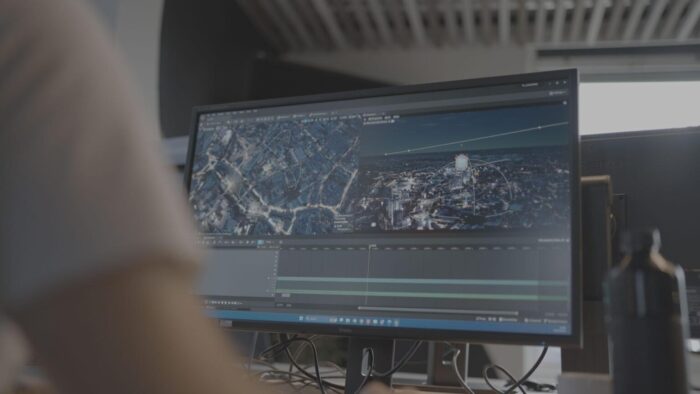 A person works at a desk with a computer monitor displaying video editing software and aerial city footage.