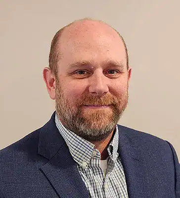 A man with a beard and short hair wearing a blue suit jacket and a checked shirt, standing in front of a neutral background.