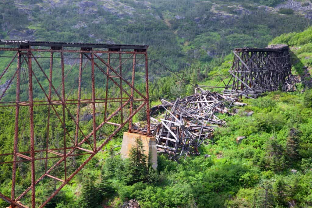 Collapsed wooden bridge structure lies between two standing steel bridge supports amid dense green forest, a poignant snapshot for any global contest on infrastructure challenges.