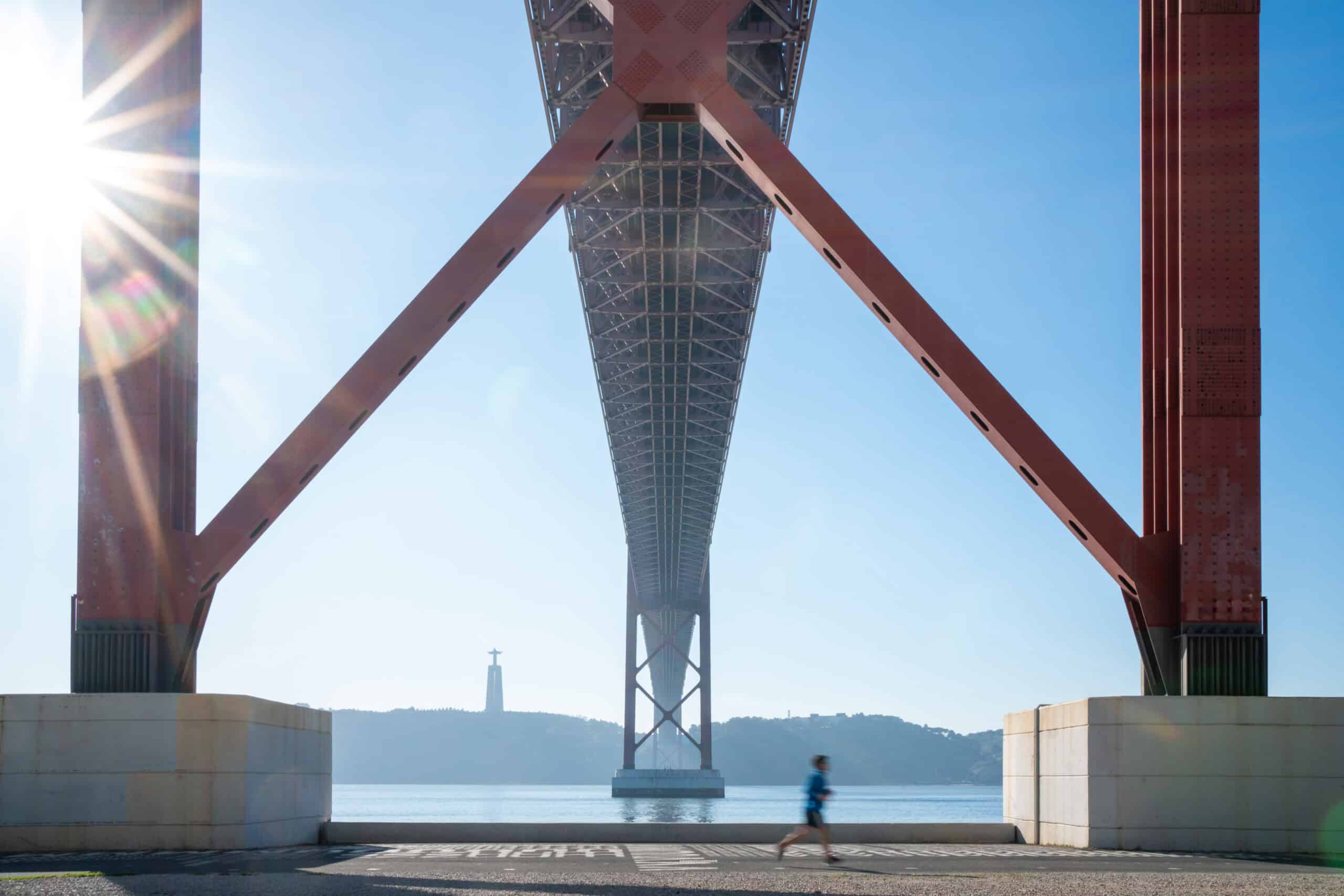 View from beneath a large red bridge with a person jogging on the pavement in the foreground and a statue visible across the water in the distance under a bright, sunny sky.