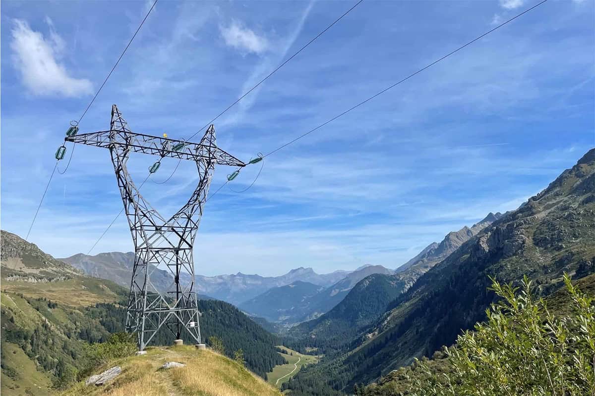 A tall electrical transmission tower, part of America's Power Grid, stands on a grassy hill overlooking a valley with mountains and a blue sky in the background.