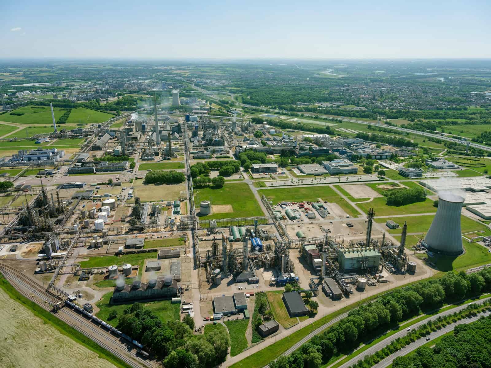 Aerial view of an industrial complex with factories, storage tanks, and a cooling tower—showcasing advanced plant production—surrounded by green fields and roads under a clear sky.