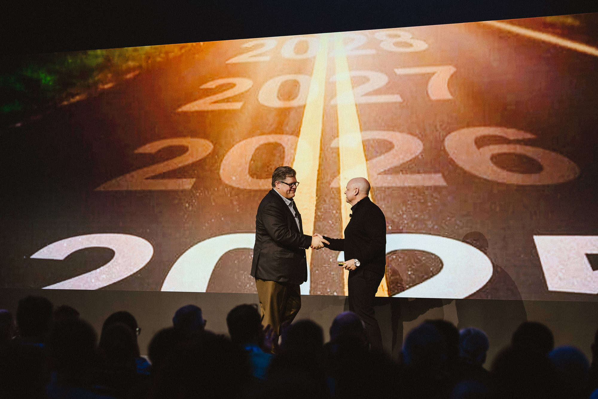Two men in suits shake hands on stage in front of a large screen displaying a road with years 2025 to 2028 marked ahead.