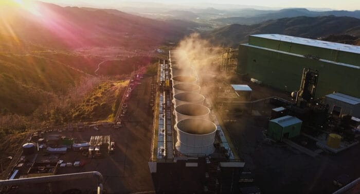 Aerial view of a power plant with cooling towers emitting steam, set against a mountainous landscape under a setting sun.