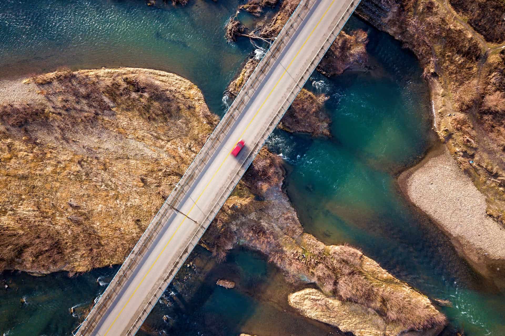 Aerial view of a red Bentley driving over a bridge spanning a river with rocky banks and patches of vegetation. The brief moment captures the elegance in motion against nature's rugged backdrop.