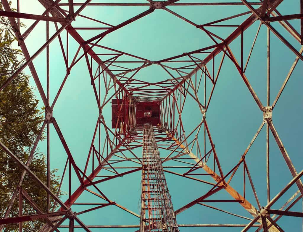 Looking straight up through the metal framework of a tall transmission tower against a clear blue sky, with a tree visible on the left side.