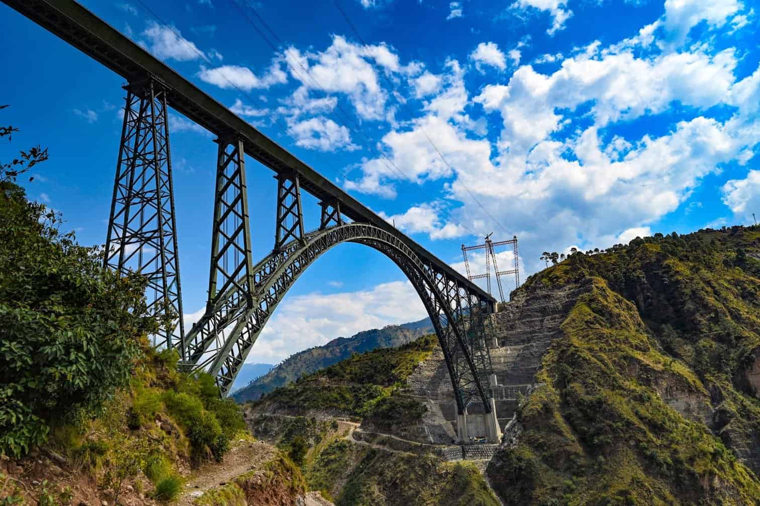 Steel arch bridge spanning a deep valley with rugged hills, under a clear blue sky with scattered clouds.