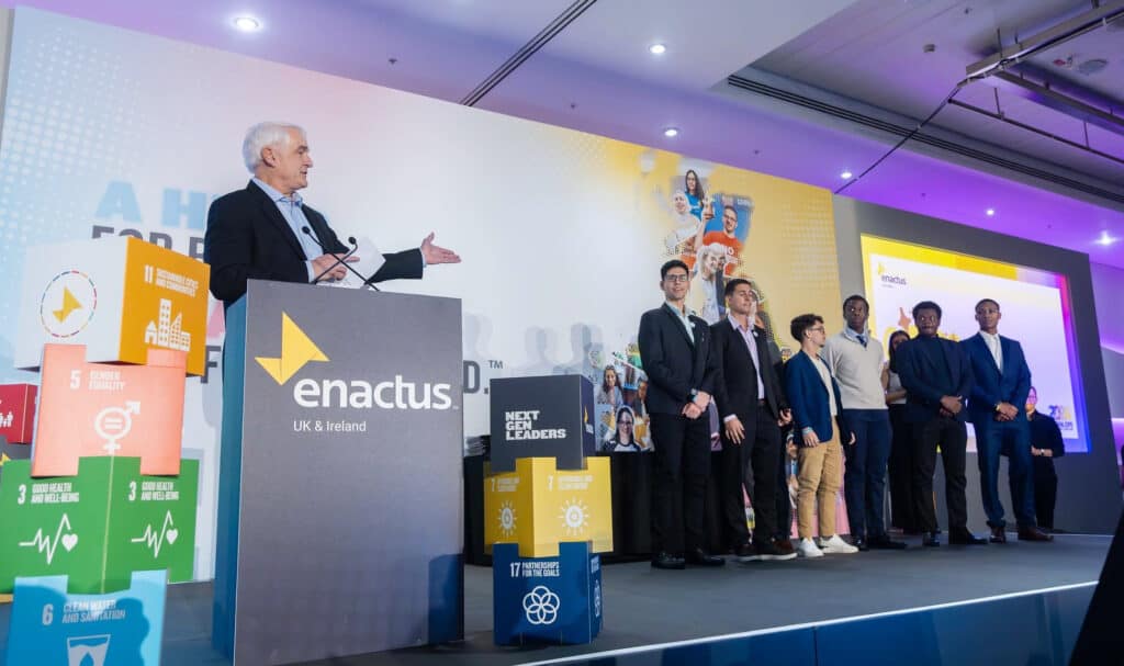 A man speaks at a podium labeled "Enactus UK & Ireland" while a group of people stand on stage beside colorful SDG-themed boxes.