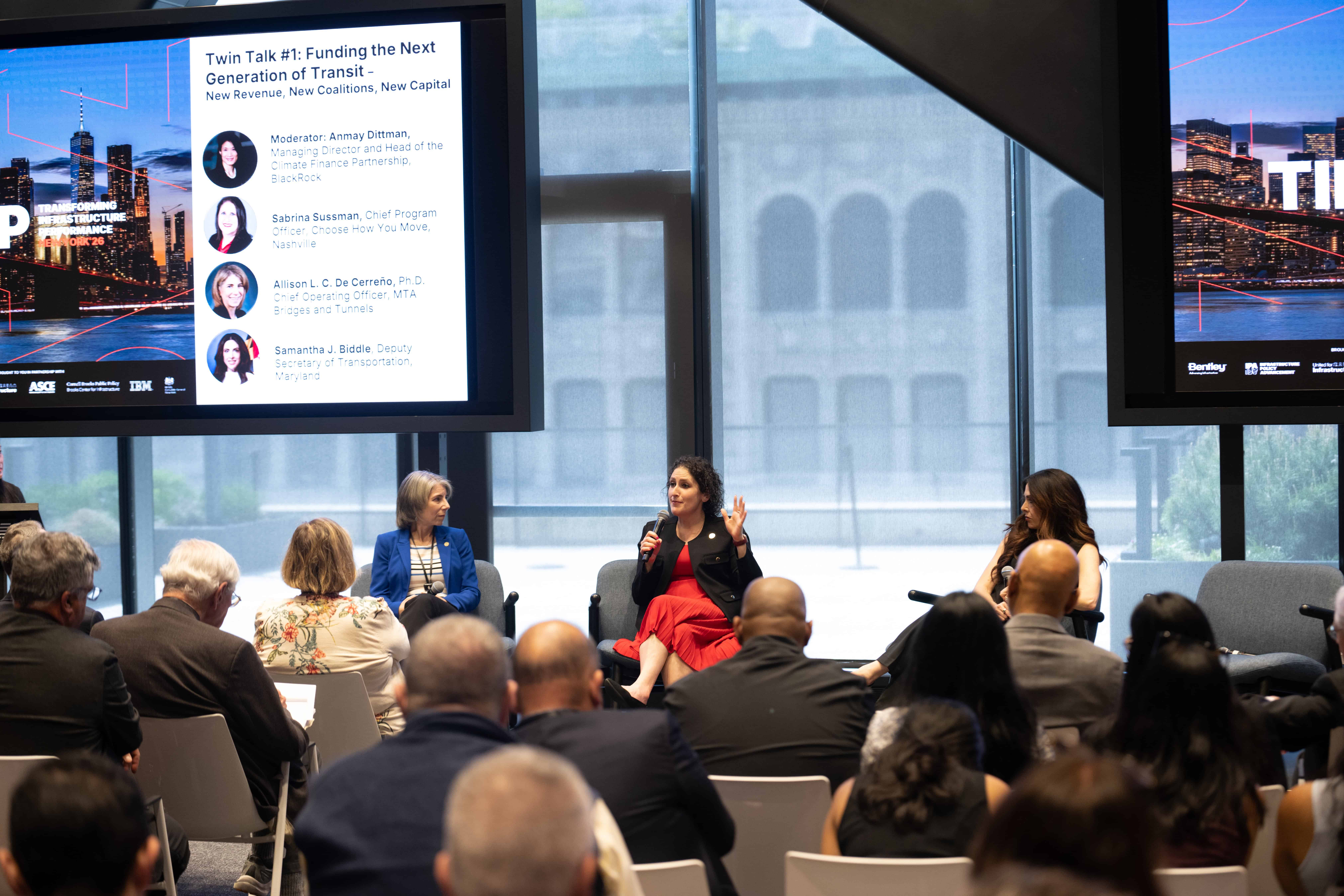 Panelists speak on stage at a U.S. Infrastructure transit funding event as audience members listen; a screen displays session information and speakers' names.