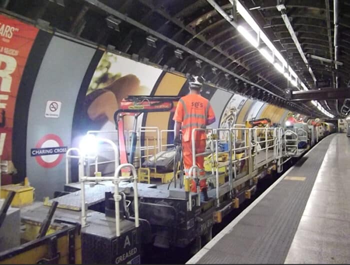 A worker in high-visibility clothing stands on maintenance equipment on a subway track at Charing Cross station. The platform is empty, with advertising posters on the tunnel walls.