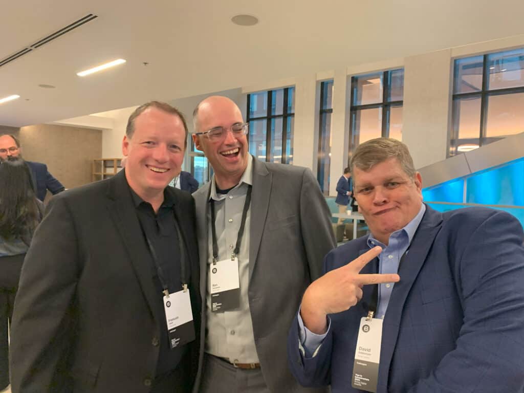 Three men in business attire pose together at an indoor event; two are smiling at the camera while one makes a peace sign gesture. All wear conference name badges.