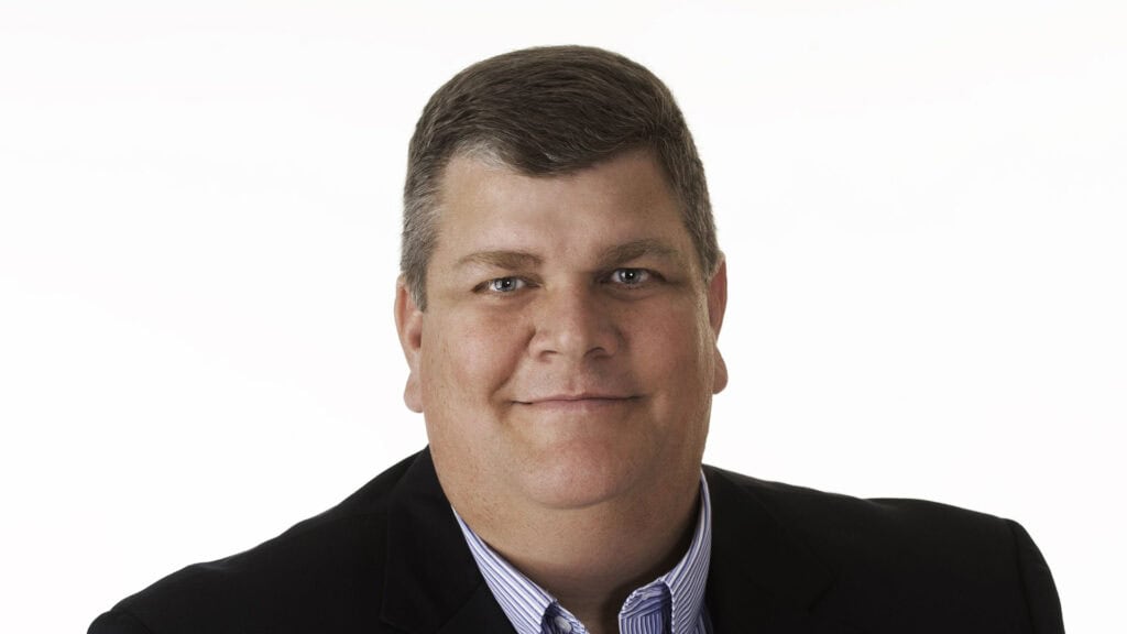 A man with short brown hair wearing a dark blazer and a light blue striped shirt, posing against a plain white background.