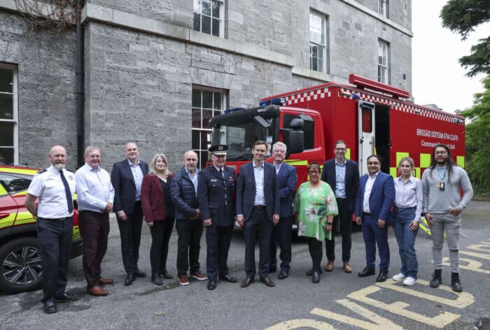 A group of people, including uniformed personnel, standing in front of a fire truck and a fire chief vehicle outside a stone building.