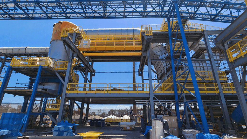 Large industrial facility with yellow railings, blue steel supports, and extensive ductwork under a clear blue sky. Equipment and materials are visible on the ground.