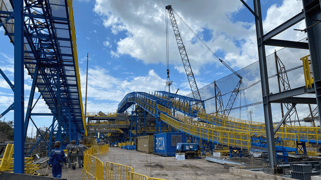 Construction site with workers, scaffolding, cranes, and yellow safety barriers under a partly cloudy sky. Central structure appears to be an industrial conveyor system.