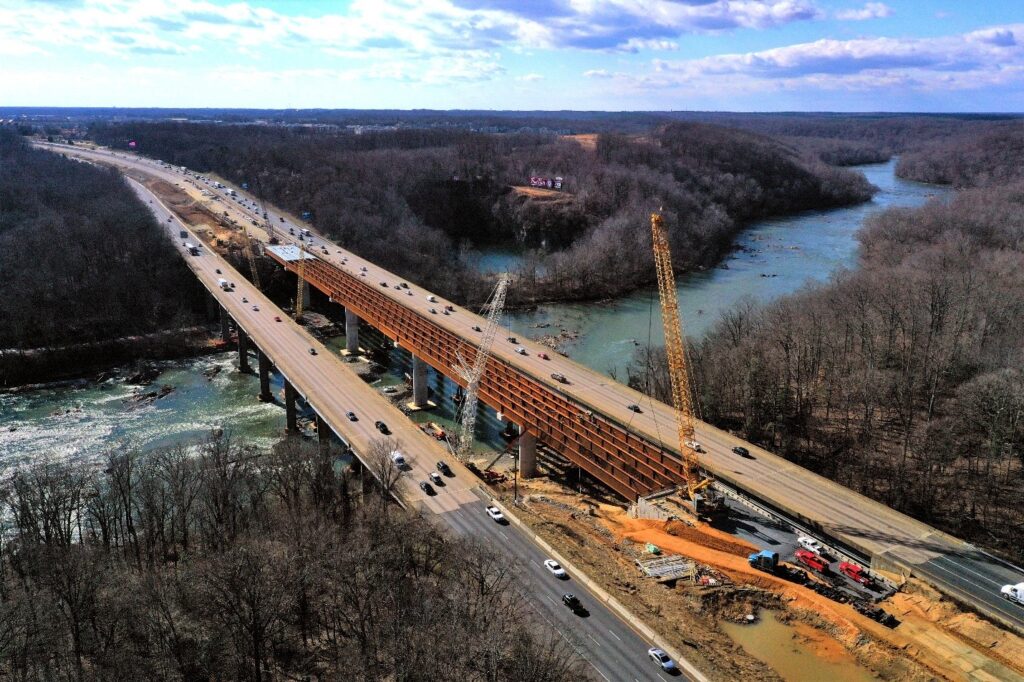 Aerial view of a highway bridge under construction over a river, showcasing cranes, construction vehicles, and ongoing traffic on adjacent lanes—highlighting modern design and innovative construction techniques.