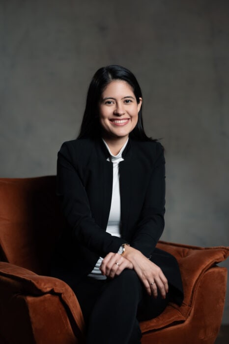 A woman with long dark hair, wearing a black blazer and white top, sits on a brown chair, smiling at the camera against a plain, neutral background.