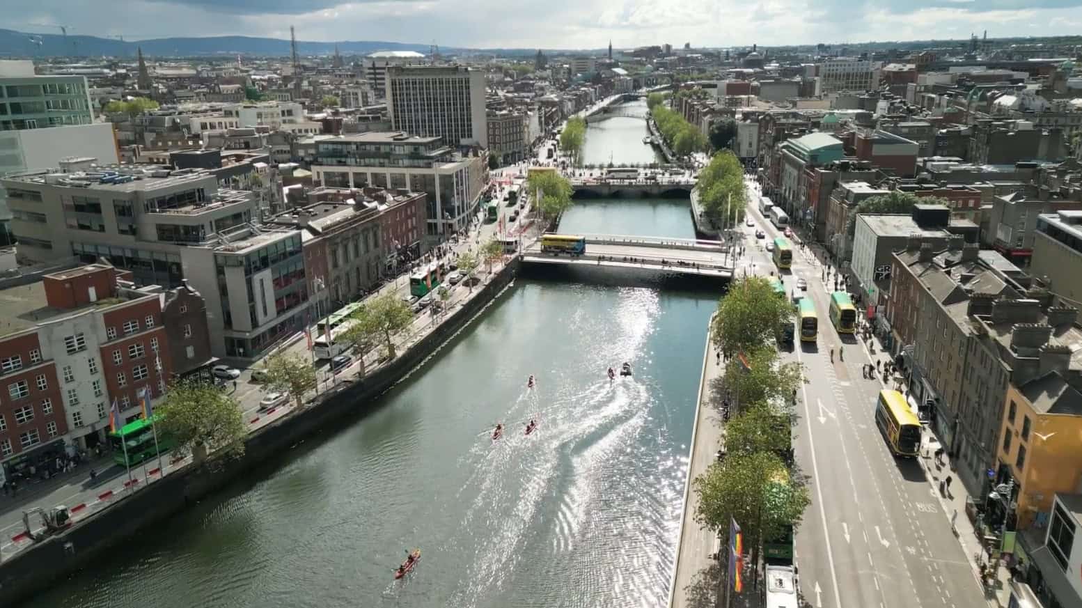 Aerial view of a city with a river running through it, bridges crossing the water, people rowing boats, and busy roads lined with buildings and trees on both sides.