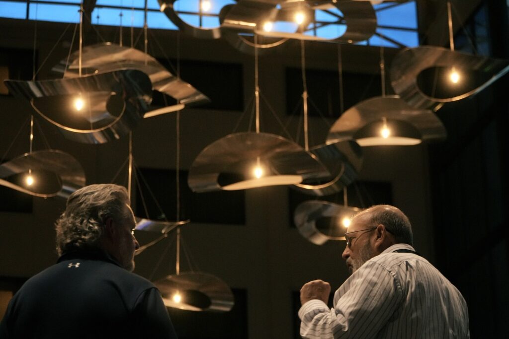 Two men stand indoors under modern, abstract metal light fixtures hanging from the ceiling, illuminated by several exposed bulbs.