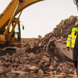 Three construction workers in high-visibility jackets and helmets walk through a rocky construction site with heavy machinery in the background.