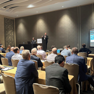 A group of people seated in a conference room listens to a presenter at the podium, with a presentation projected on screen during the AEC Fall Conference 2025