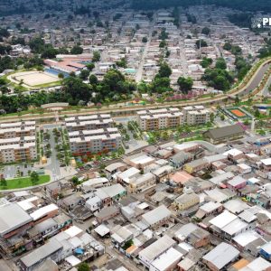 Aerial view of a densely populated urban area with new residential buildings and green spaces under development, labeled "PROSAMIN+" and "Visão Geral.