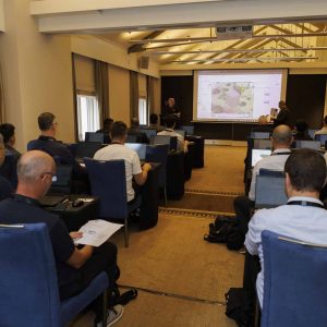 People seated in a conference room with laptops, watching a presenter speak at the front near a screen displaying a map.