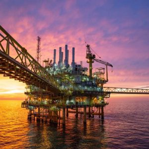 An offshore oil platform stands in the ocean at sunset, showcasing its infrastructure journey with industrial structures, cranes, and a gas flare silhouetted against a colorful sky.