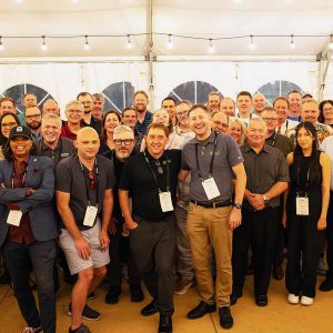 A large group of people pose and smile for a group photo inside a tent, many wearing name badges, under string lights at a Bentley Master Classes event.
