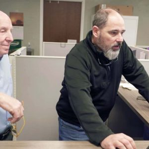Two men stand at a desk in an office, looking at a computer monitor displaying OpenSite+; one types while the other observes, collaborating on site design.