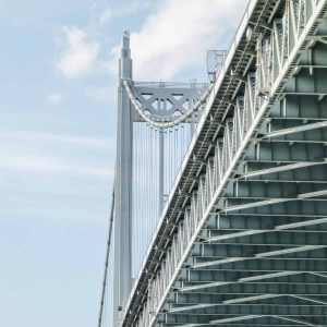 Close-up view of a suspension bridge’s steel framework and cables against a blue sky with some clouds.