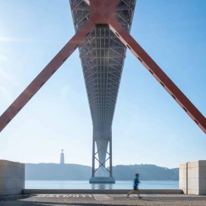 View from beneath a large red bridge with a person jogging on the pavement in the foreground and a statue visible across the water in the distance under a bright, sunny sky.