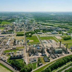 Aerial view of an industrial complex with factories, storage tanks, and a cooling tower—showcasing advanced plant production—surrounded by green fields and roads under a clear sky.