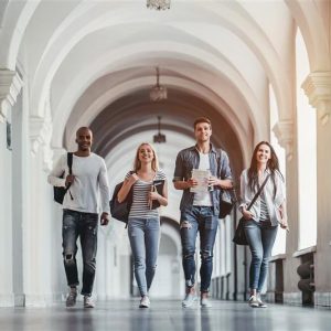 Four Gen Z young adults casually walk down a bright, arched hallway, each carrying bags or books, dressed in casual attire, and appearing relaxed.