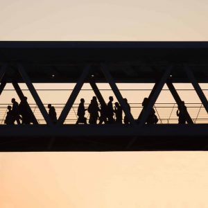 Silhouettes of people walking across an enclosed pedestrian bridge at sunset evoke the quiet transformation brought by AI disruption and technology infrastructure shaping modern city life.