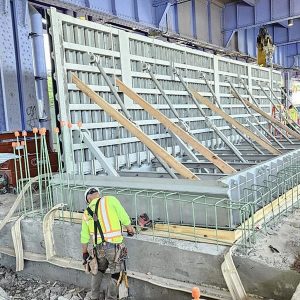 Construction workers in NYC reinforce a concrete structure under a large metal bridge, using wooden supports and metal rebar. Tools and equipment around the site highlight efforts toward climate resilience.