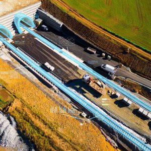 Aerial view of a tunnel construction site with two tunnel entrances, construction vehicles, and digital graphics overlaid on the image.