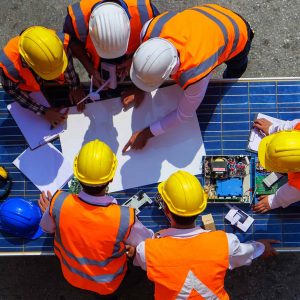 Construction workers in high visibility vests and safety helmets discussing plans over a blueprint with a solar panel nearby.