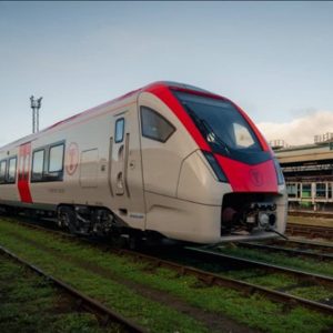 A modern passenger train with red and white livery is stationary on railway tracks beside an industrial building under a partly cloudy sky.