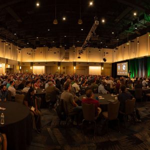 The 2025 Bentley Civil User Conference features a large conference room filled with attendees at round tables, all focused on the stage where multiple screens showcase the latest engineering insights and presentations.