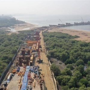 Aerial view of a coastal construction site with heavy machinery surrounded by greenery, a sandy beach, and a pier extending into the sea.