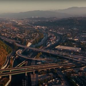Aerial view of multiple highways intersecting in an urban area, showcasing impressive infrastructure design, surrounded by houses, hills, and cityscape in the background at sunset.