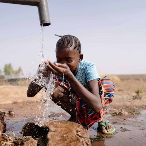 A child crouches by a pipe, drinking water with hands cupped—an image of hope and resilience in an outdoor, rural setting with dry grass and scattered trees, showing the power of survival on our changing planet.