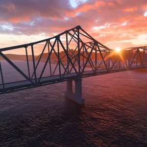 A steel truss bridge, expertly crafted using Bridge Design principles, crosses a wide river at sunset, with the sun partially visible near the horizon and clouds scattered across the sky.