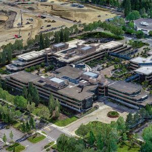 Aerial view of a large office complex surrounded by trees, parking lots, and a nearby construction site using Intelligent Digital Twins for unified reality modeling and efficient earth-moving equipment deployment.