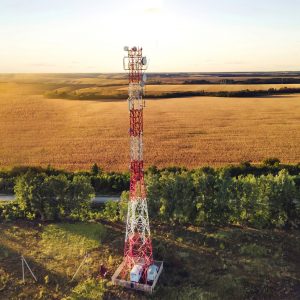 A red and white telecommunications tower stands in front of a large golden field with trees and distant farmland under a clear sky—Know Your Tower and its vital role in connecting rural landscapes.