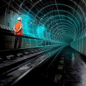 A person in a hard hat and safety vest stands on railroad tracks in a tunnel with digital grid lines and data network overlays.