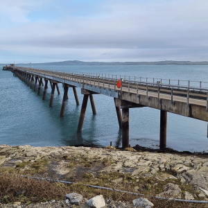 A long concrete pier, part of the Holyhead Jetty inspection by Royal HaskoningDHV, extends over calm blue water, with a rocky shore in the foreground and distant land visible on the horizon under a cloudy sky. A long concrete pier, part of the Holyhead Jetty inspection by Royal HaskoningDHV, extends over calm blue water, with a rocky shore in the foreground and distant land visible on the horizon under a cloudy sky.
